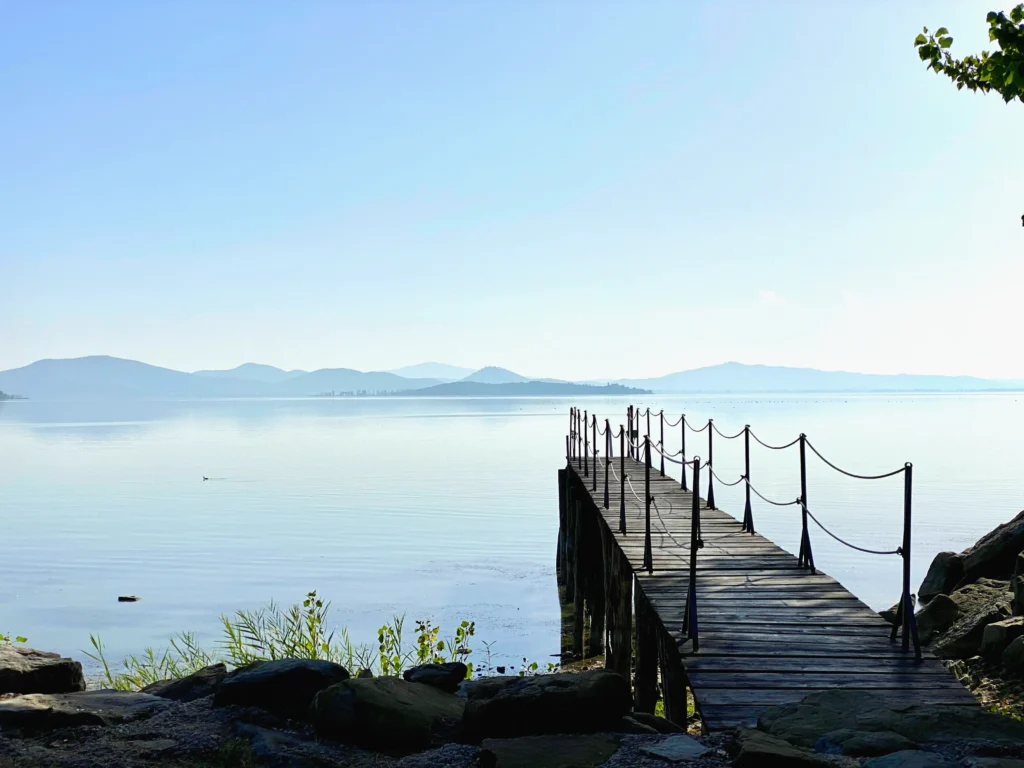 Holzsteg mit Seilgeländer führt hinaus auf einen ruhigen See, am Horizont Hügel im Morgendunst